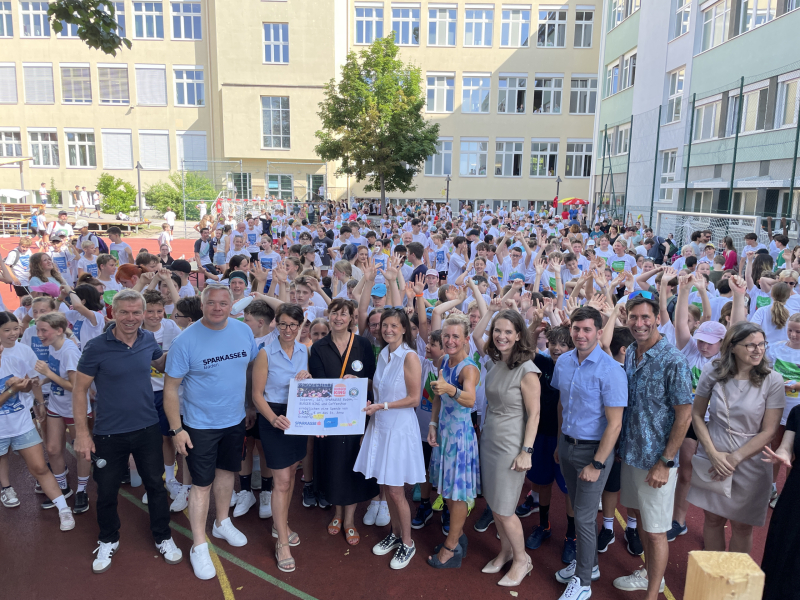 Hunderte Schülerinnen strecken ihre Hände in die Höhe. Im Vordergrund steht eine Reihe von Sponsoren bei der Scheckübergaabe an die Pflegedirektorin des St. Anna Kinderspitals