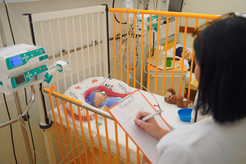 Eine Übungspuppe liegt im Patientinnenbett. Im Vordergrund macht eine Übungsteilnehmende Notizen.