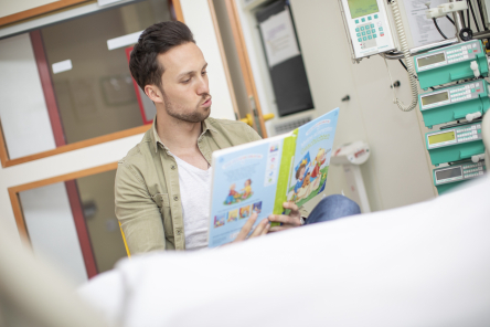 Mann liest aus einem Buch an einem Krankenbett.